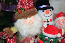 A group of festive decorations featuring Santa Claus figures and a snowman. The central Santa has a plush beard, red hat with white patterns, and green holly leaves with red berries. To the right, a snowman with a black top hat, orange nose, and striped scarf is smiling. Another Santa figure, more detailed and plastic-like, appears alongside a whimsical green character with oversized eyes and a Santa hat. Christmas tree branches are partially visible in the background.