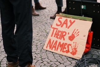 A cardboard sign with the words 'SAY THEIR NAMES' painted in red, accompanied by two red handprints. The sign is supported against a speaker or amplifier on a cobblestone ground, with partially visible legs of people in the background.