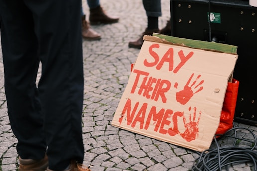 A cardboard sign with the words 'SAY THEIR NAMES' painted in red, accompanied by two red handprints. The sign is supported against a speaker or amplifier on a cobblestone ground, with partially visible legs of people in the background.