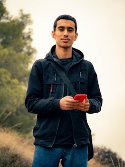 A person stands outdoors holding a red phone, wearing a black jacket and blue jeans. The background features blurred greenery and dry grass.