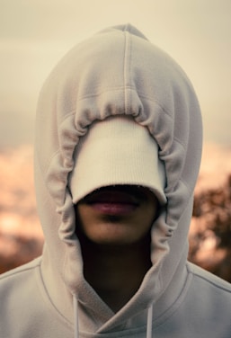 A person with their hood pulled over their face, obscuring their eyes and partially covering their nose and mouth. The fabric of the hoodie is a light color, providing a soft and casual appearance. The background is blurred with warm tones.