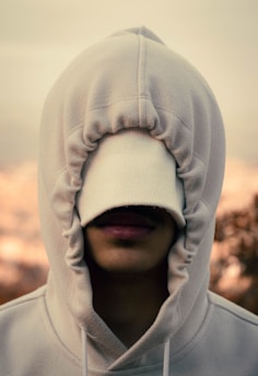 A person with their hood pulled over their face, obscuring their eyes and partially covering their nose and mouth. The fabric of the hoodie is a light color, providing a soft and casual appearance. The background is blurred with warm tones.