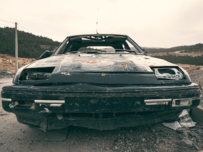 A dilapidated and rusted car sits abandoned on a rough terrain, surrounded by a sparsely vegetated landscape with distant hills. The car's front end is damaged, with missing headlights and a battered bumper. The paint is severely worn and peeling, indicating long-term neglect.