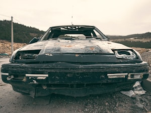 A dilapidated and rusted car sits abandoned on a rough terrain, surrounded by a sparsely vegetated landscape with distant hills. The car's front end is damaged, with missing headlights and a battered bumper. The paint is severely worn and peeling, indicating long-term neglect.