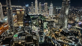 A vibrant cityscape at night featuring high-rise buildings and skyscrapers illuminated with lights. The architecture includes modern designs with reflective glass surfaces. Streets are bustling with traffic, and the area is densely built, showcasing the urban lifestyle.