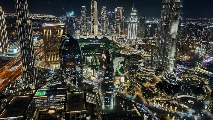 A vibrant cityscape at night featuring high-rise buildings and skyscrapers illuminated with lights. The architecture includes modern designs with reflective glass surfaces. Streets are bustling with traffic, and the area is densely built, showcasing the urban lifestyle.