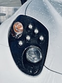 A close-up of a car headlight featuring multiple circular light elements arranged in a stylish design. The surface is covered in water droplets, suggesting the car has recently been washed or is in the rain. The headlight is set against a sleek, shiny surface.