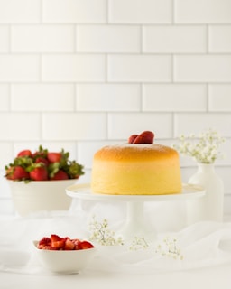 A fluffy, golden-brown cake sits on a white pedestal with fresh strawberries on top. In the background, a white bowl filled with strawberries and a simple white vase with small white flowers add elegance. The setting exudes a clean and minimalistic aesthetic with a soft white backdrop tiled wall.