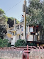 A Brutalist architectural structure with concrete slabs, geometric shapes, and red accents. The building is partially obscured by trees and surrounded by a red metal fence. The design features a variety of angular components, creating a complex and textured facade.