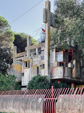A Brutalist architectural structure with concrete slabs, geometric shapes, and red accents. The building is partially obscured by trees and surrounded by a red metal fence. The design features a variety of angular components, creating a complex and textured facade.