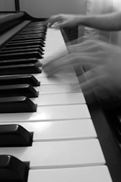 Blurred hands playing the keys of a piano, suggesting movement and speed. The piano keys are prominently displayed in black and white.