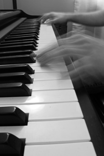 Blurred hands playing the keys of a piano, suggesting movement and speed. The piano keys are prominently displayed in black and white.