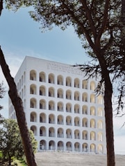 A large, white, classical building with multiple rows of arched windows is framed by two trees in the foreground. The structure reflects a blend of modernist and ancient Roman architectural styles. The building's facade has inscriptions at the top. The sky is clear and blue, adding to the serene setting.