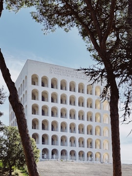 A large, white, classical building with multiple rows of arched windows is framed by two trees in the foreground. The structure reflects a blend of modernist and ancient Roman architectural styles. The building's facade has inscriptions at the top. The sky is clear and blue, adding to the serene setting.