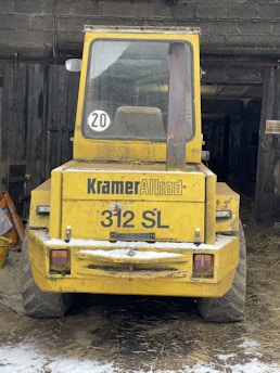 A yellow industrial vehicle is parked in a barn-like structure with its rear facing the viewer. The vehicle is labeled 'Kramer Allrad 312 SL' and has a number '20' sticker in the window. The vehicle is surrounded by some snow and hay on the ground, and there are wooden beams and a dark interior visible in the background.