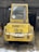 A yellow industrial vehicle is parked in a barn-like structure with its rear facing the viewer. The vehicle is labeled 'Kramer Allrad 312 SL' and has a number '20' sticker in the window. The vehicle is surrounded by some snow and hay on the ground, and there are wooden beams and a dark interior visible in the background.