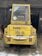 A yellow industrial vehicle is parked in a barn-like structure with its rear facing the viewer. The vehicle is labeled 'Kramer Allrad 312 SL' and has a number '20' sticker in the window. The vehicle is surrounded by some snow and hay on the ground, and there are wooden beams and a dark interior visible in the background.