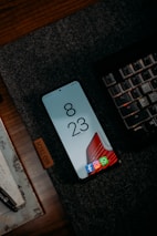 A smartphone is placed on a dark textured mat beside a mechanical keyboard. The phone screen displays the time 8:23 with several app icons at the bottom such as Facebook, Instagram, and WhatsApp. There is a wooden surface beneath the mat and a portion of a pen and a piece of paper can also be seen.