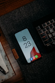A smartphone is placed on a dark textured mat beside a mechanical keyboard. The phone screen displays the time 8:23 with several app icons at the bottom such as Facebook, Instagram, and WhatsApp. There is a wooden surface beneath the mat and a portion of a pen and a piece of paper can also be seen.
