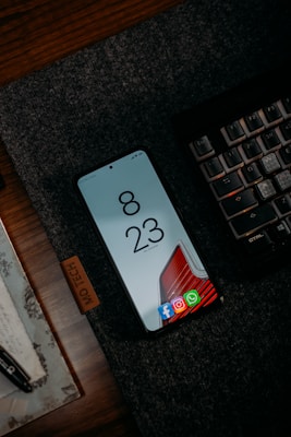 A smartphone is placed on a dark textured mat beside a mechanical keyboard. The phone screen displays the time 8:23 with several app icons at the bottom such as Facebook, Instagram, and WhatsApp. There is a wooden surface beneath the mat and a portion of a pen and a piece of paper can also be seen.