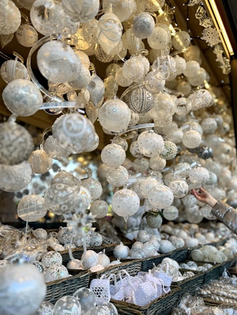 An array of intricately designed white ornaments, predominantly spherical, hang from above, while more are piled in wicker baskets below. Delicate patterns and textures adorn each piece, giving a sense of craftsmanship and elegance. A hand reaches in from the right, likely selecting an ornament from the display. Soft lighting highlights the sparkle and shine of the ornaments, enhancing their festive appearance.