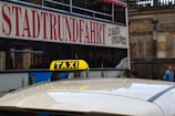 A taxi with a yellow roof sign is situated in front of a double-decker tour bus bearing the word 'STADTRUNDFAHRT' on its side. The background features a stone building facade and two pedestrians walking nearby.