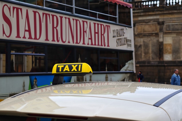 A taxi with a yellow roof sign is situated in front of a double-decker tour bus bearing the word 'STADTRUNDFAHRT' on its side. The background features a stone building facade and two pedestrians walking nearby.