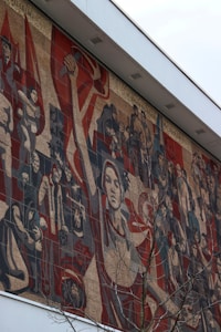 A large mosaic mural depicting a variety of figures engaged in dynamic activities, likely representing themes of labor and unity. The figures are styled in a socialist realism manner, with strong and determined expressions. The central figure is holding a hammer, symbolizing industrial work. The mural conveys a sense of movement and strength, with a prominent use of reds and earth tones.