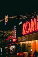A busy street scene at night featuring brightly lit buildings with neon signs. The word 'Komedia' glows in red, and festive lights are strung across the street. Decorative elements include a large pair of red and white striped legs and a circular light fixture shaped like an owl. People walk below the illuminated signage.