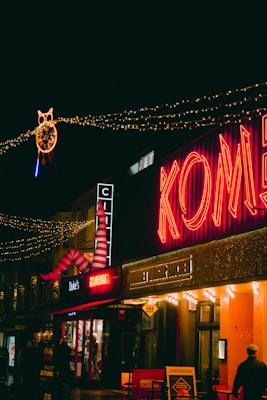 A busy street scene at night featuring brightly lit buildings with neon signs. The word 'Komedia' glows in red, and festive lights are strung across the street. Decorative elements include a large pair of red and white striped legs and a circular light fixture shaped like an owl. People walk below the illuminated signage.