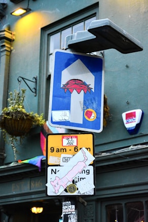 Two street signs are seen, one above the other, both with multiple stickers, including ones depicting cartoon-like characters and various designs. The background includes a building with a window, a wall-mounted plant basket, and an additional sign.