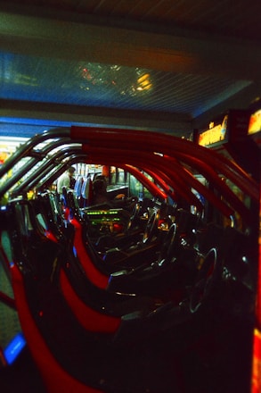 A row of arcade racing game machines with vibrant red frames and steering wheels lined up in a dimly lit gaming area. The environment includes neon lights and reflections contributing to a retro, nostalgic atmosphere. A person is visible in the background, potentially engaged in one of the machines.