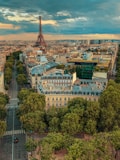 An aerial view of Paris, showcasing the Eiffel Tower in the background. The city is filled with historic buildings, tree-lined streets, and a vibrant urban landscape. The sky is partly cloudy, adding a dramatic effect to the scene.