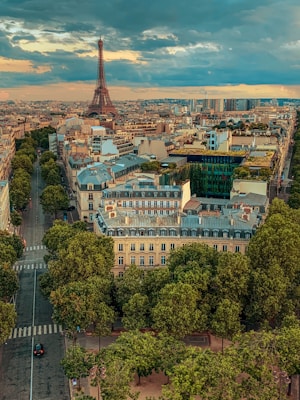An aerial view of Paris, showcasing the Eiffel Tower in the background. The city is filled with historic buildings, tree-lined streets, and a vibrant urban landscape. The sky is partly cloudy, adding a dramatic effect to the scene.