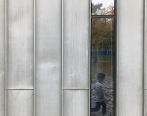 A reflective surface with vertical panels shows a blurred reflection of a child walking on a patterned path. The background features a park with trees displaying autumn foliage in various yellow hues.