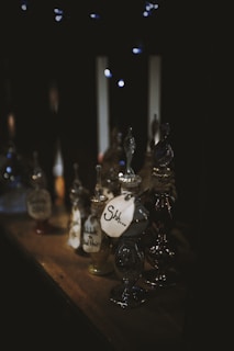 A collection of intricately designed glass bottles placed on a wooden shelf. Each bottle has a decorative stopper and a label with mysterious writing, including one labeled 'Shh...' The scene is dimly lit with a warm, cozy ambiance complemented by faint twinkling lights in the background.