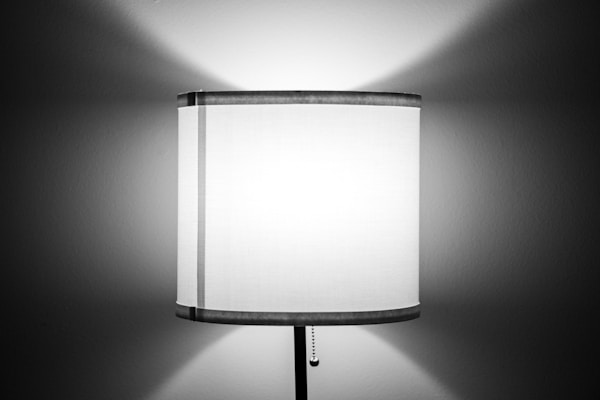 A cylindrical lampshade with a pull chain hangs from a lamp, casting a symmetrical spread of light onto a plain wall. The fabric of the shade is light in color, allowing light to softly diffuse through it.