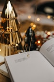 An open book with poetic text is placed on a surface. A delicate necklace with a crescent moon pendant hangs above the book. In the background, there are decorative elements including golden and black conical objects that resemble trees, and blurred warm lights create a cozy atmosphere.