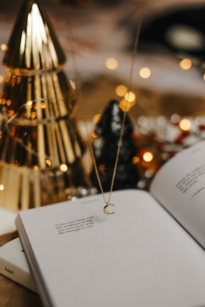 An open book with poetic text is placed on a surface. A delicate necklace with a crescent moon pendant hangs above the book. In the background, there are decorative elements including golden and black conical objects that resemble trees, and blurred warm lights create a cozy atmosphere.