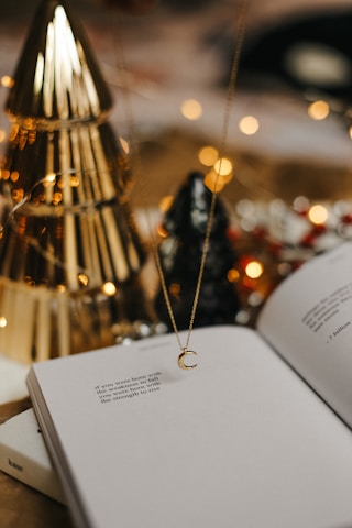 An open book with poetic text is placed on a surface. A delicate necklace with a crescent moon pendant hangs above the book. In the background, there are decorative elements including golden and black conical objects that resemble trees, and blurred warm lights create a cozy atmosphere.