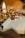 A small toy figure with glasses and a wand stands on an open book. The book's page reads 'Chapter One: The Boy Who Lived.' Soft, warm lighting creates a cozy atmosphere, with golden and silver decorations, including a triangular object, visible in the blurred background.