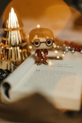 A small toy figure with glasses and a wand stands on an open book. The book's page reads 'Chapter One: The Boy Who Lived.' Soft, warm lighting creates a cozy atmosphere, with golden and silver decorations, including a triangular object, visible in the blurred background.