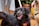 A black dog with a happy expression wearing a colorful bandana is the focal point. Its tongue is out, and it seems to be on a leash. Another dog is partially visible, and there are humans around, possibly walking the dogs. The setting appears to be outdoors with plants and a wooden deck.