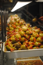 A large tray filled with skewered green olives wrapped in slices of red cured meat, displayed on a counter under bright lighting. The arrangement appears abundant and artisanal, likely in a market or deli setting.