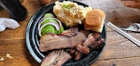 A black plate on a wooden table holds slices of smoked brisket, garnished with pickles, red onion, and cucumber, along with a serving of creamy potato salad and a small bun. Cutlery and a straw are placed nearby.