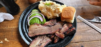 A black plate on a wooden table holds slices of smoked brisket, garnished with pickles, red onion, and cucumber, along with a serving of creamy potato salad and a small bun. Cutlery and a straw are placed nearby.
