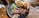 A black plate on a wooden table holds slices of smoked brisket, garnished with pickles, red onion, and cucumber, along with a serving of creamy potato salad and a small bun. Cutlery and a straw are placed nearby.