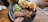 A black plate on a wooden table holds slices of smoked brisket, garnished with pickles, red onion, and cucumber, along with a serving of creamy potato salad and a small bun. Cutlery and a straw are placed nearby.