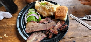 A black plate on a wooden table holds slices of smoked brisket, garnished with pickles, red onion, and cucumber, along with a serving of creamy potato salad and a small bun. Cutlery and a straw are placed nearby.