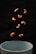 Several cashew nuts are captured in mid-air against a dark background, appearing to be falling towards a white bowl positioned at the bottom of the image.